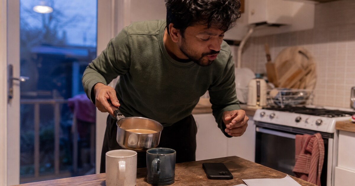 A South Asian man in his early thirties with short wavy hair, wearing a faded green jumper and loose dark trousers, leans towards a phone…