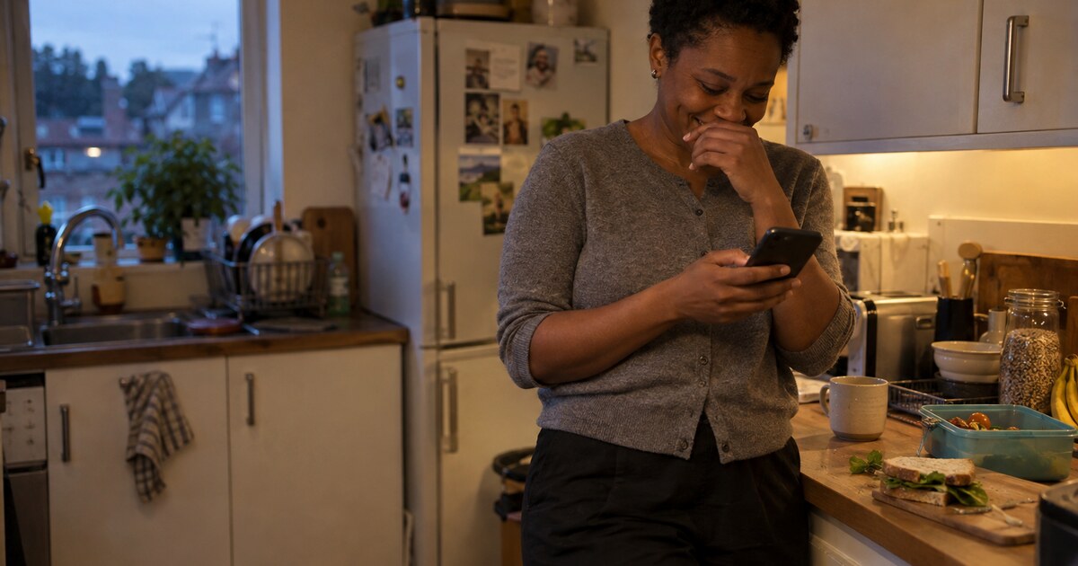 A Black woman in her mid-thirties with short natural hair, wearing a plain grey cardigan and dark trousers, stands against the counter in a…