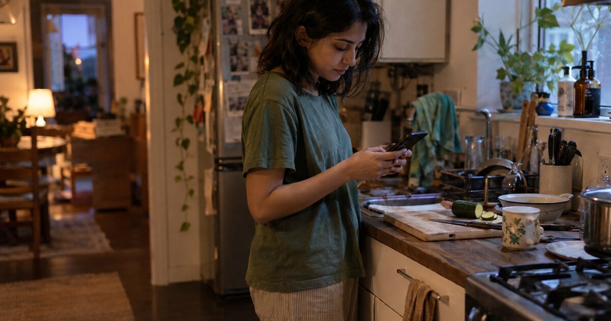 A South Asian woman in her late twenties with shoulder-length dark hair, wearing a faded green T-shirt and loose cotton trousers, stands at…
