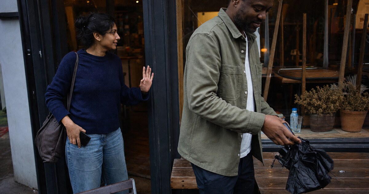 A South Asian woman in her early thirties wearing a navy jumper and worn jeans stands outside a small neighbourhood café at early evening,…
