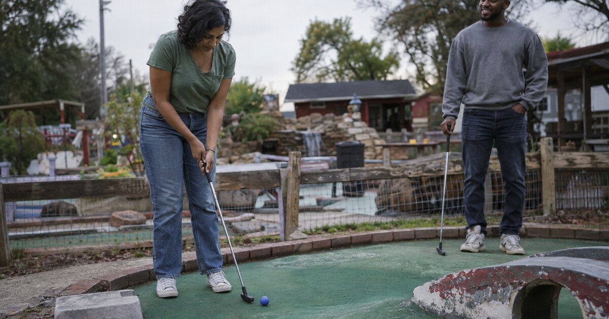 At a small neighbourhood mini golf course in the late afternoon, a South Asian woman in her early thirties with short curly hair, a faded…