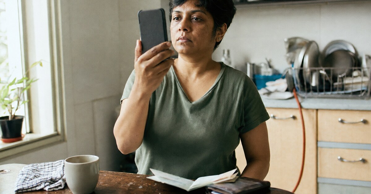 A South Asian woman in her late thirties with short dark hair, wearing a faded green T-shirt, stands beside a scratched kitchen table in…