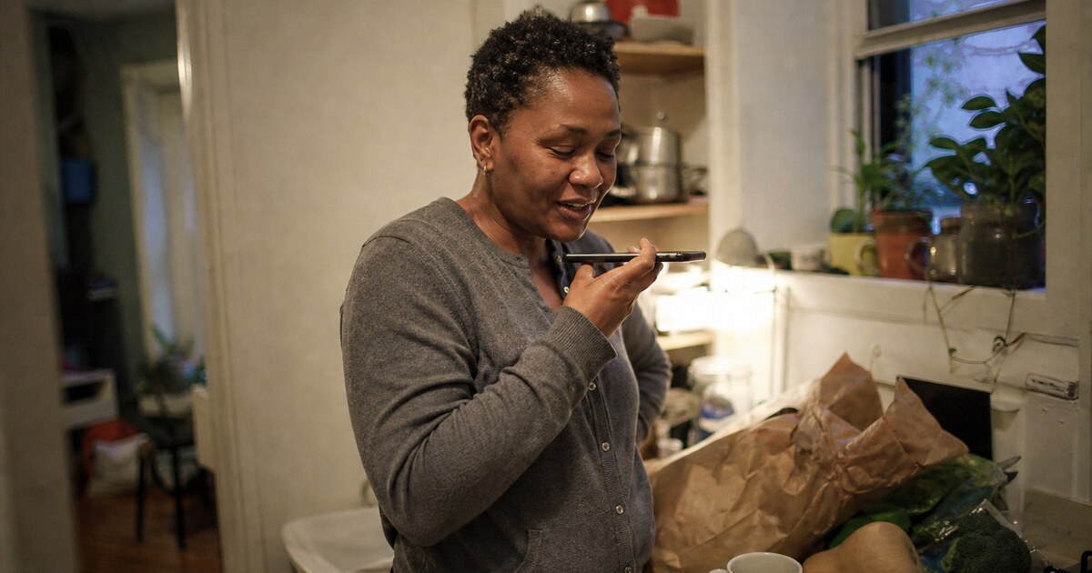 A Black woman in her late forties with short natural hair, wearing a faded grey cardigan and dark trousers, stands beside a cluttered…